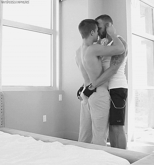 Two men embrace in a passionate kiss. The larger man firmly grasps the other man’s rear, while the smaller of the two removes the other’s earbuds.