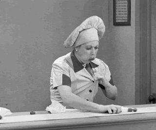 A woman is standing in front of a conveyer belt in a chocolate factory, scooping up every chocolate that passes and shoving them down her top.