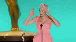 Hannah Waddingham dressed for an awards ceremony (pink, tight, long gown) is on stage accepting a win, and doing a 'mind blown' action with her hands.