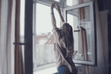 A woman stands in front of open windows. The light linen curtains are flapping around her, and her top is waving in the breeze. She has her arms raised above her head.