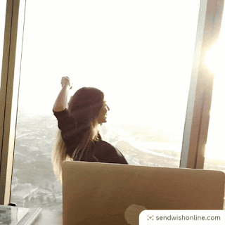 A woman is sitting on an office swivel chair, at a desk with a computer on it. Behind her is a beautiful skyline view of a cityscape. The sun is setting, it's stunning. She spins on her swivel chair, raising her arms in celebration, and turns away, momentarily, from her screen.