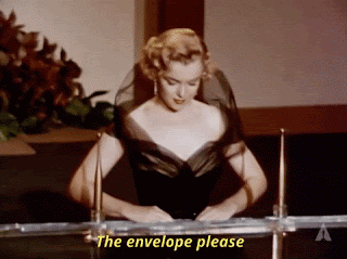 Marilyn Monroe at an awards ceremony. SHe's at the podium in a posh, black, off the shoulder dress, and the caption says, 'The envelope please.' Then someone off the camera, hands her an envelope.