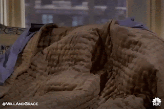 Debra Messing, as Grace from Will and Grace, is under the bed covers. She lifts them up so her face is revealed. She looks dishevelled, confused, and like she needs to go back to sleep.