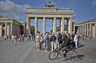The Brandenburg gate on a sunny, clear-skyed day. There's a huddle of tourists milling around and a guy on a bike, circling in the foreground.