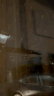 The view from my front door when the snow started. It's constant, and coming down regularly, and illuminated in the light outside my front door. In the background, there's another house, and the side of a car on the driveway is visible.