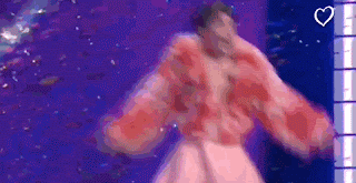 Nemo, a person with curly brown hair, wearing pink and red puffy sleeves, walks onstage, with their arms outstretched. Ther face suggests they are emotional and elated.