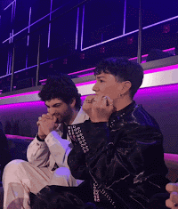 A young caucasian brunette man is sitting in a greenroom area, reacting to being given 12 points. His face moves from disbelief to joy, and he stands up and sits down in his seat while moving his arms in the air. It's a spontaneous reaction to a thrill.