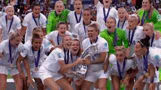 The England Women's football team, in their kit, with the Euros trophy, lifting it as one in celebration.