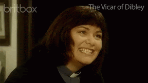 Dawn French, as the vicar of Dibley, is wearing a black shirt and white dog collar. She's taking to someone off camera, and goes a wide grin with teeth bared - giving a really sarcastic smile.