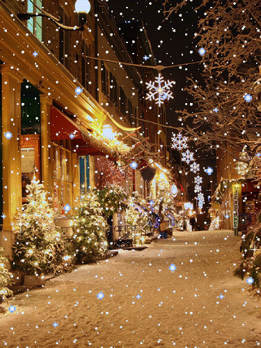 This may contain: a snowy street with christmas lights and snowflakes on the trees in front of buildings