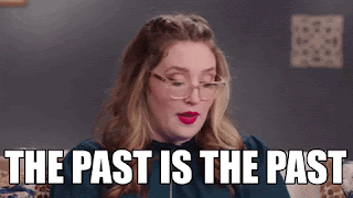 A white woman with blonde hair and round glasses is talking flippantly and saying, 'The past is the past.'