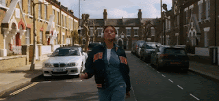 A young woman, walks down a residential street, with headphones. She walks with a swagger.