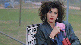 Marisa Tomei in My Cousin Vinny, is sitting on a park bench, holding a pink camera on an angle, and casually taking a photo without much thought.