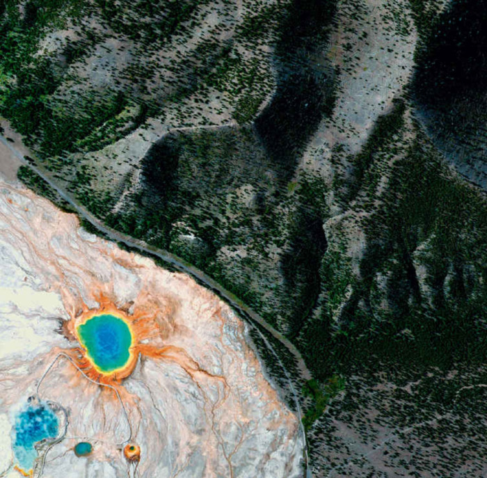green mountains with trees and circular hot spring atop orange and grey rock. the spring is made up of concentric circles which look rainbow.